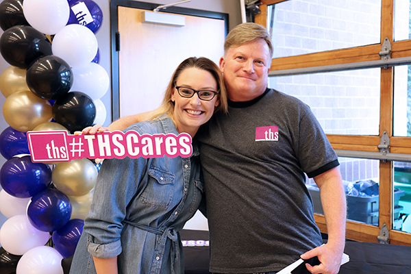 two employees giving a side hug while holding a #THSCares sign representing positive careers