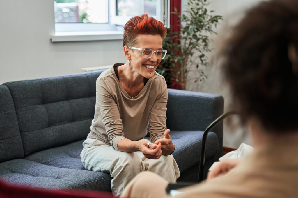 Woman laughing out loud while sitting at sofa and talking with her doctor during therapy A woman with short red hair is smiling as she leans over toward their counselor.