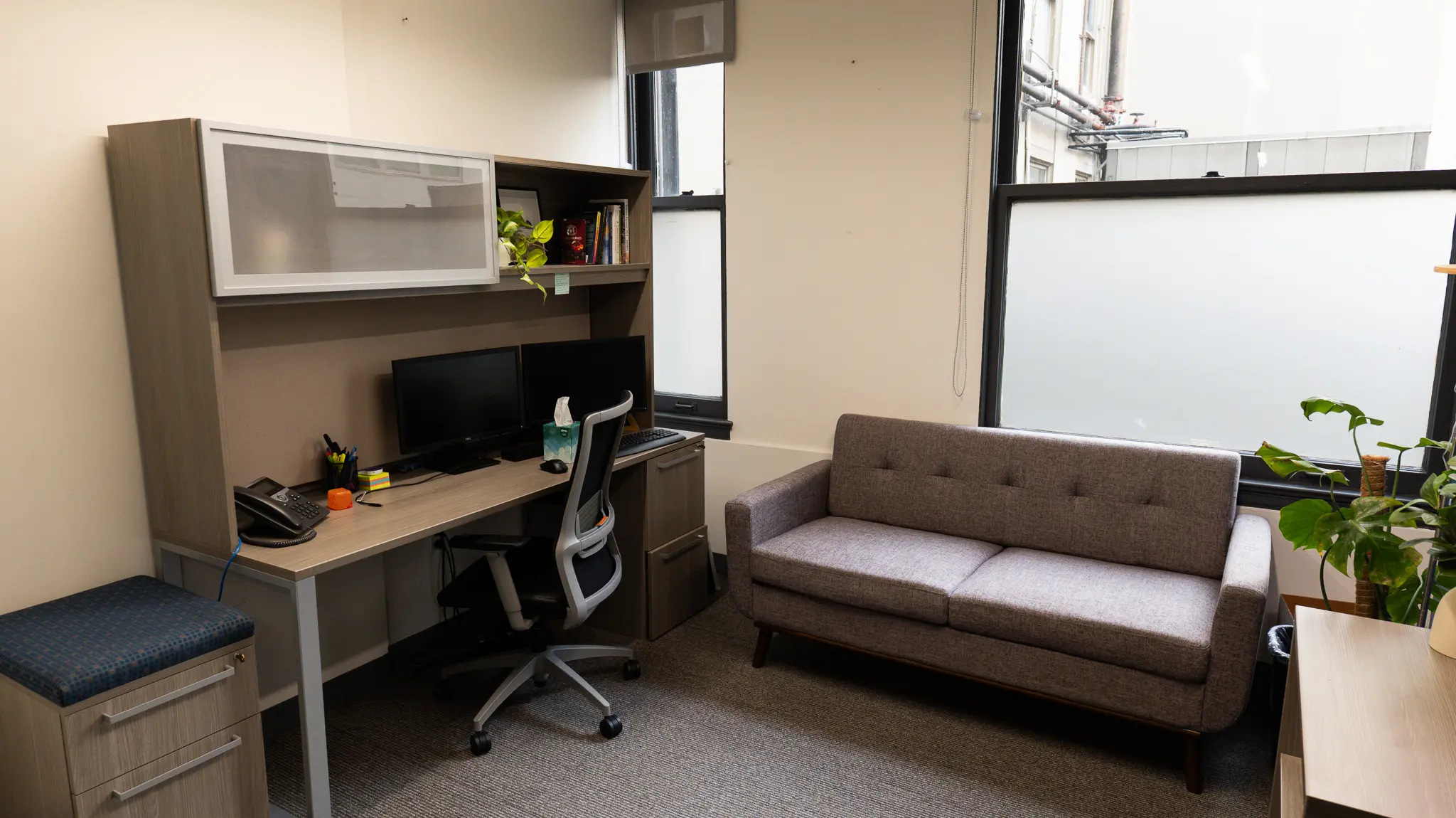 ths-olive-branch-location_office1 Private therapy office at THS Olive Branch featuring a counselor's desk, bookshelf with plants, and a gray tufted loveseat for client sessions