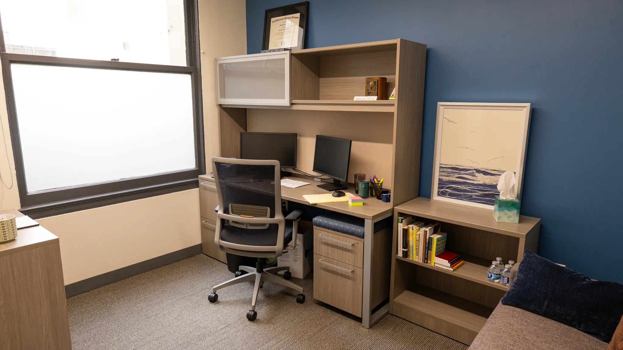 ths-olive-branch-location_office2 THS Olive Branch therapist office with blue accent wall, wood desk, dual monitors, framed credentials, and a small bookshelf with a tissue box