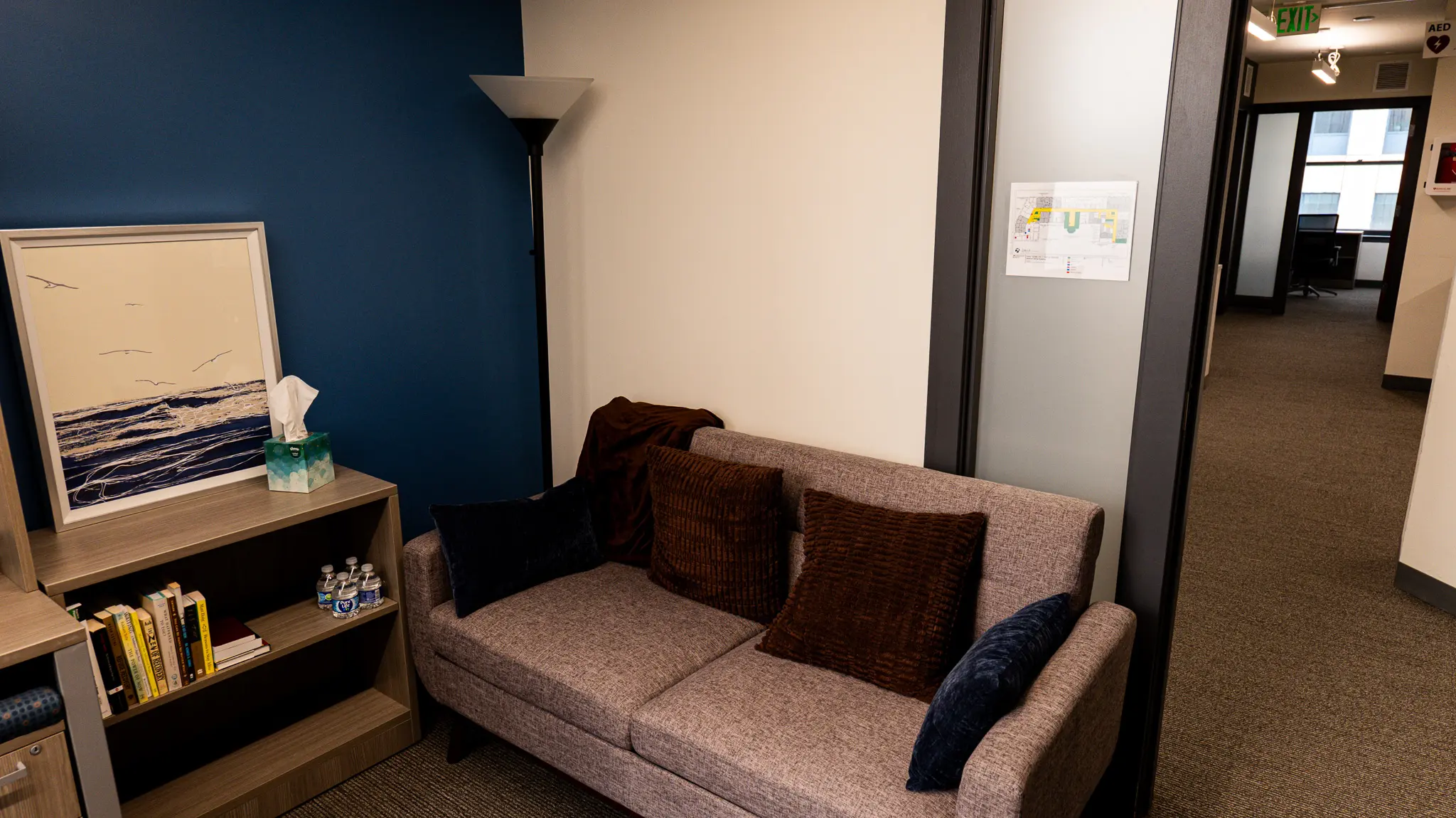 ths-olive-branch-location_office4 THS Olive Branch counseling office with gray loveseat, throw pillows, bookshelf, ocean artwork, and a view down the office hallway