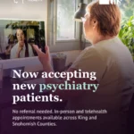 Photo of an older woman seen from behind, sitting at a desk near a sunlit window with green plants, gesturing while talking to a provider visible on her computer screen during a video call. Purple gradient rises from the bottom of the image. Mental Health badge (head with heart-and-cross icon) top left, THS logo top right. Bold white and mint serif headline: "Now accepting new psychiatry patients." Subtext notes no referral is needed and appointments are available in-person and via telehealth across King and Snohomish Counties. Bottom left, mint text reads "Ready to talk to someone?" followed by "1-833-278-HELP · Link in bio." Mint phone icon bottom right.